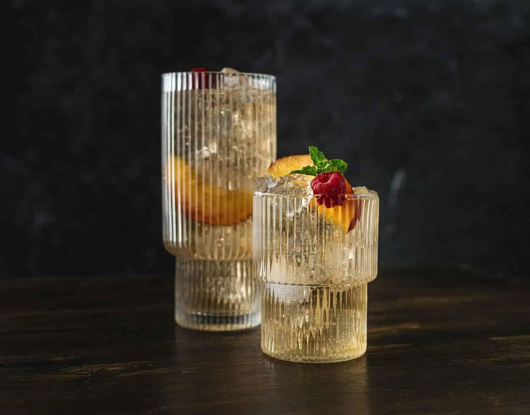 Two cocktails with ice, peach slices, raspberry, and mint in ribbed glasses on a dark table