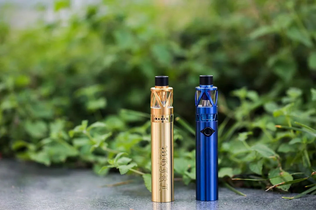 Gold and blue disposable weed vapes on outdoor surface with green foliage background