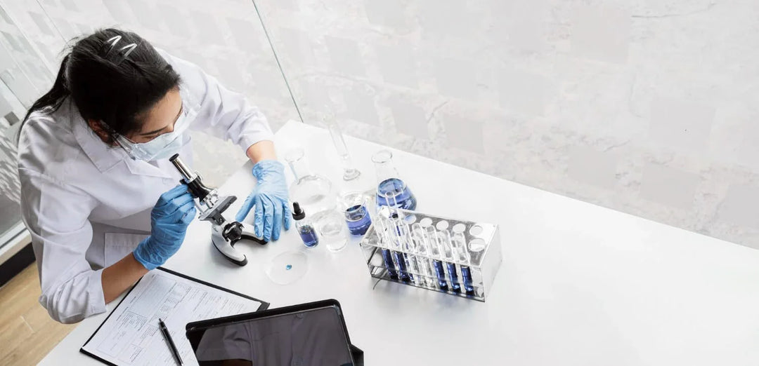Scientist in lab coat using microscope with blue liquid samples and test tubes on table