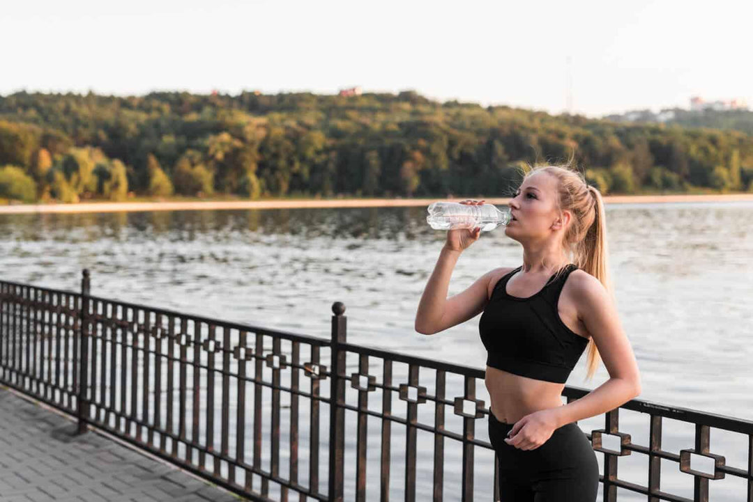 Woman in athletic wear drinking water outdoors by a lake after exercise