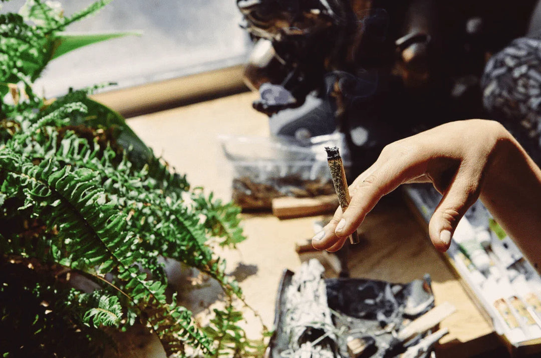 Hand holding a lit pre-rolled cone joint by a table with green fern and rolling supplies