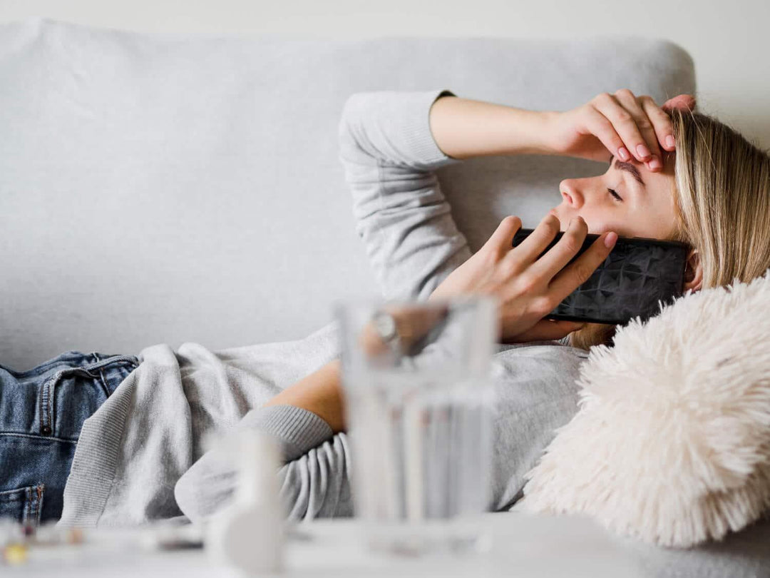 Woman lying on couch feeling unwell, talking on phone, medicine and water on table