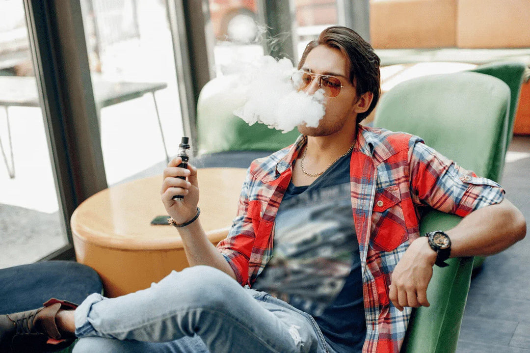 Young man vaping indoors, relaxed on a chair, wearing sunglasses and casual clothes