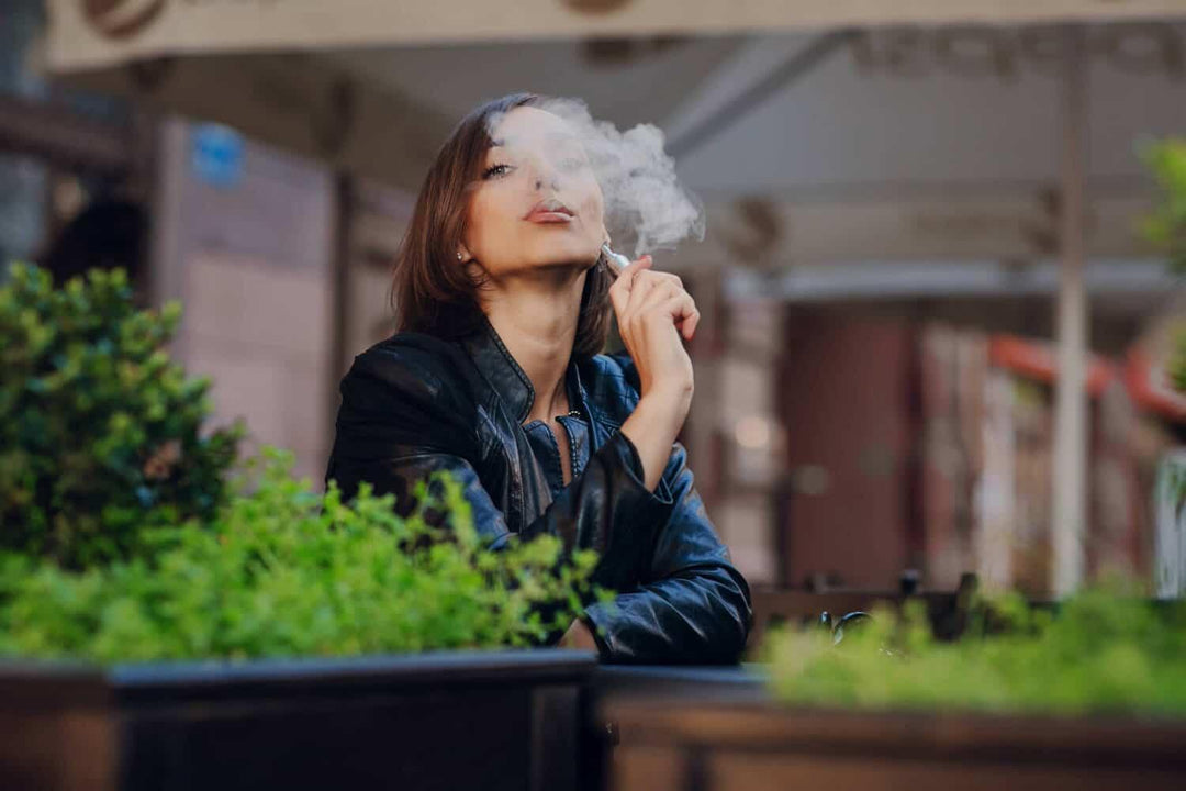 Woman vaping outdoors at a café, surrounded by plants, wearing a black leather jacket
