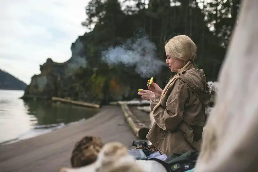 Woman vaping outdoors near a forested lakeshore, holding a yellow vape device