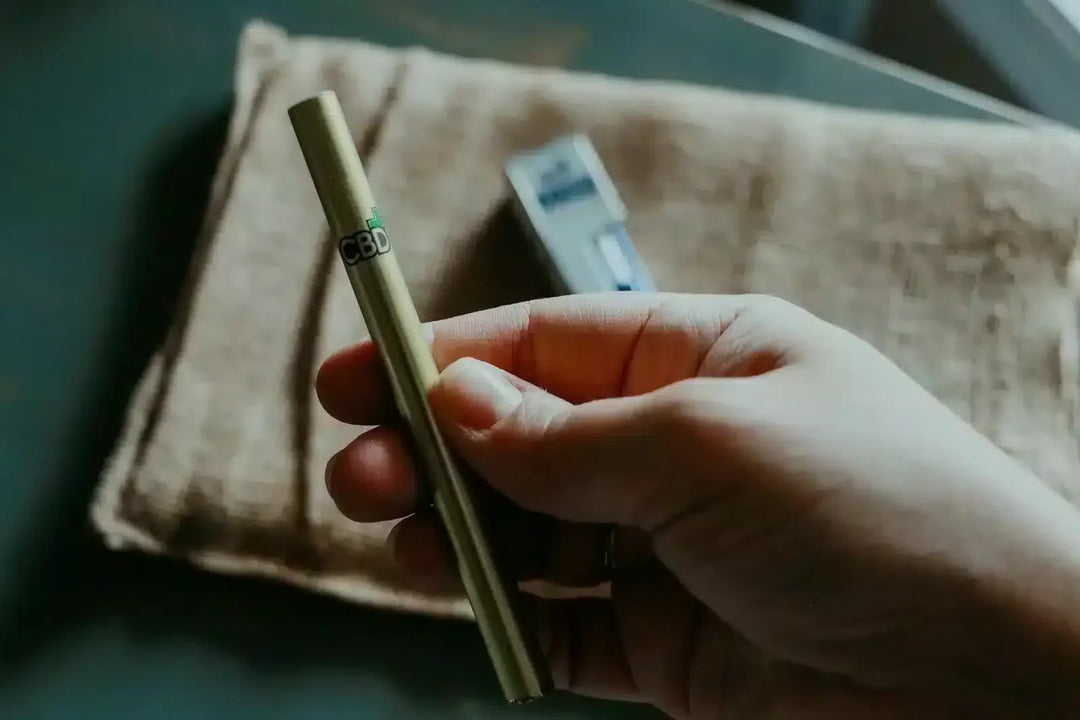 Hand holding a disposable CBD vape pen over a fabric surface, vape packaging in background