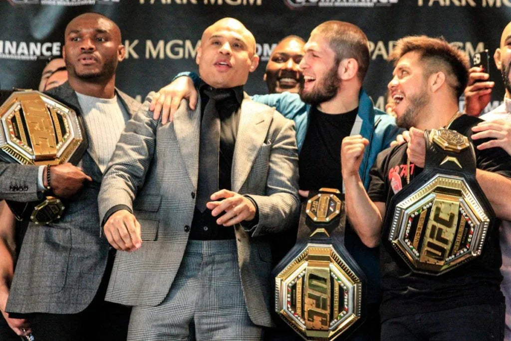 Four men celebrating at a UFC press event, three holding UFC championship belts.